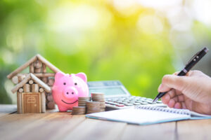 Picture of someone writing in a notebook next to two miniature houses, a piggy bank, piles of coins, and a calculator.