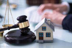 Picture of a miniature house figurine next to a gavel and the scales of justice with an attorney typing in the background.