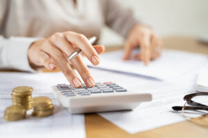 Picture of a woman holding a pen and using a calculator near glasses, papers, and stacks of coins.