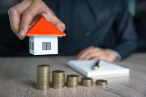 Picture of a man holding a miniature house figurine next to a pen, a stack of papers, and stacks of coins.