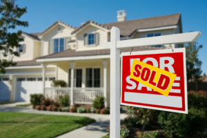 Picture of a for sale sign with a sold sticker outside of a house.