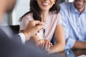 Picture of a man handing keys to a smiling woman, with another man sitting next to her.