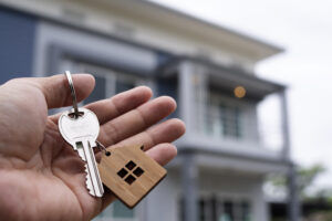 Picture of a person's hand holding a key and a house-shaped keychain with a house in the background.