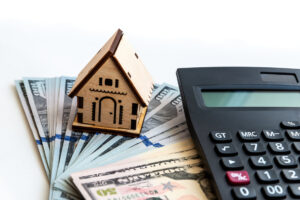 Picture of a miniature wooden house figurine, a stack of cash, and a calculator.