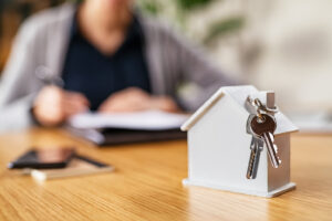 Picture of a miniature house figurine with two silver keys hanging off the chimney, with a woman signing paperwork in the background.