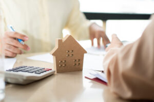 Picture of two people sitting at a table with a house figurine, a calculator, and papers between them.