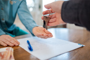 Picture of a man handing a woman keys with papers and a pen on the table between them.