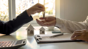 Picture of one person handing keys to another person over a table with a calculator, a clipboard, a pen, two house figurines, and a coffee cup.