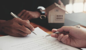 Picture of a person signing paperwork while another person points to the signature line, with a miniature house figurine to the side.