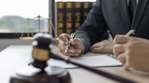 Picture of a person signing a will on a desk in between a gavel and a scales of justice figurine.