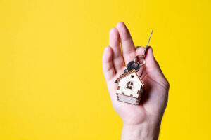 Picture of a person holding a key with a house-shaped keychain against a yellow background.