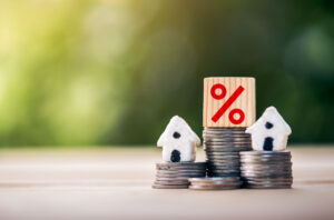 Picture of two miniature house figurines, a wooden block with a percentage sign on it, and stacks of coins.