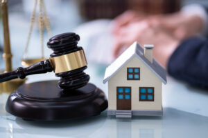 Picture of the scales of justice, a gavel, and a miniature house figurine in front of a lawyer in the background.