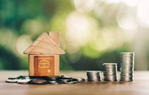 Picture of a wooden house figurine sitting on a pile of coins, with stacks of coins next to it.