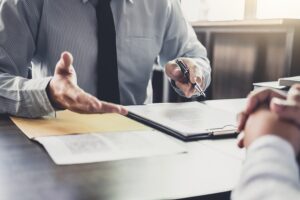 Picture of a foreclosure attorney holding a pen and gesturing to a clipboard as he talks to a client over his desk.