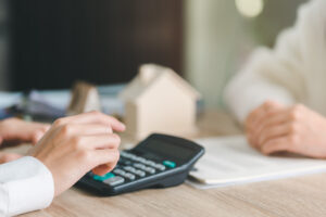 Picture of an estate planning lawyer using a calculator while speaking to a client, with papers and a wooden house figurine on the desk between them.