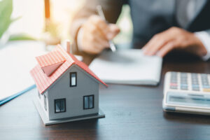 Picture of a person signing loan modification documents on a desk near a house figurine and a calculator. 