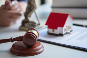 Picture of a gavel, a wooden house figurine, a clipboard with paper, and the scales of justice on a desk with a real estate attorney sitting in the background.