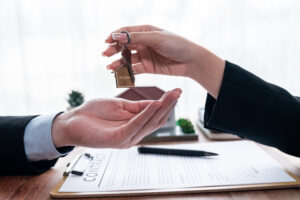 Picture of one person handing a set of house keys to another person with a pen, papers, and a clipboard in the background.