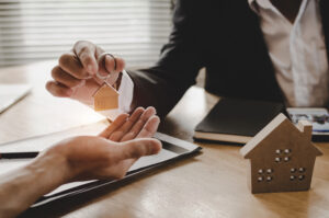 Picture of a lawyer handing a house-shaped keychain to a client over a desk, with documents and a miniature house figurine sitting on the surface.