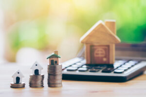 Picture of three small house figurines on top of stacks of coins, next to a larger house figurine on top of a calculator.