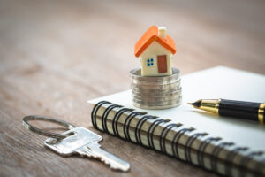 Picture of a key, a miniature house figurine, a stack of coins, a pen, and a notebook.